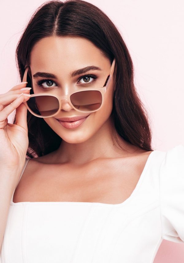 Fashion beauty portrait of young brunette woman with evening stylish  makeup and perfect clean skin. Sexy model with long curly hair posing in studio on pink background. Taking off sunglasses